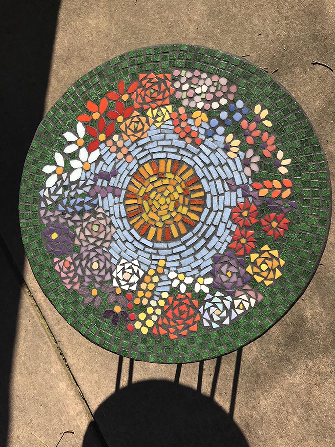 Julie Landberg's finished mosaic table with sun in sky design over field of flowers.